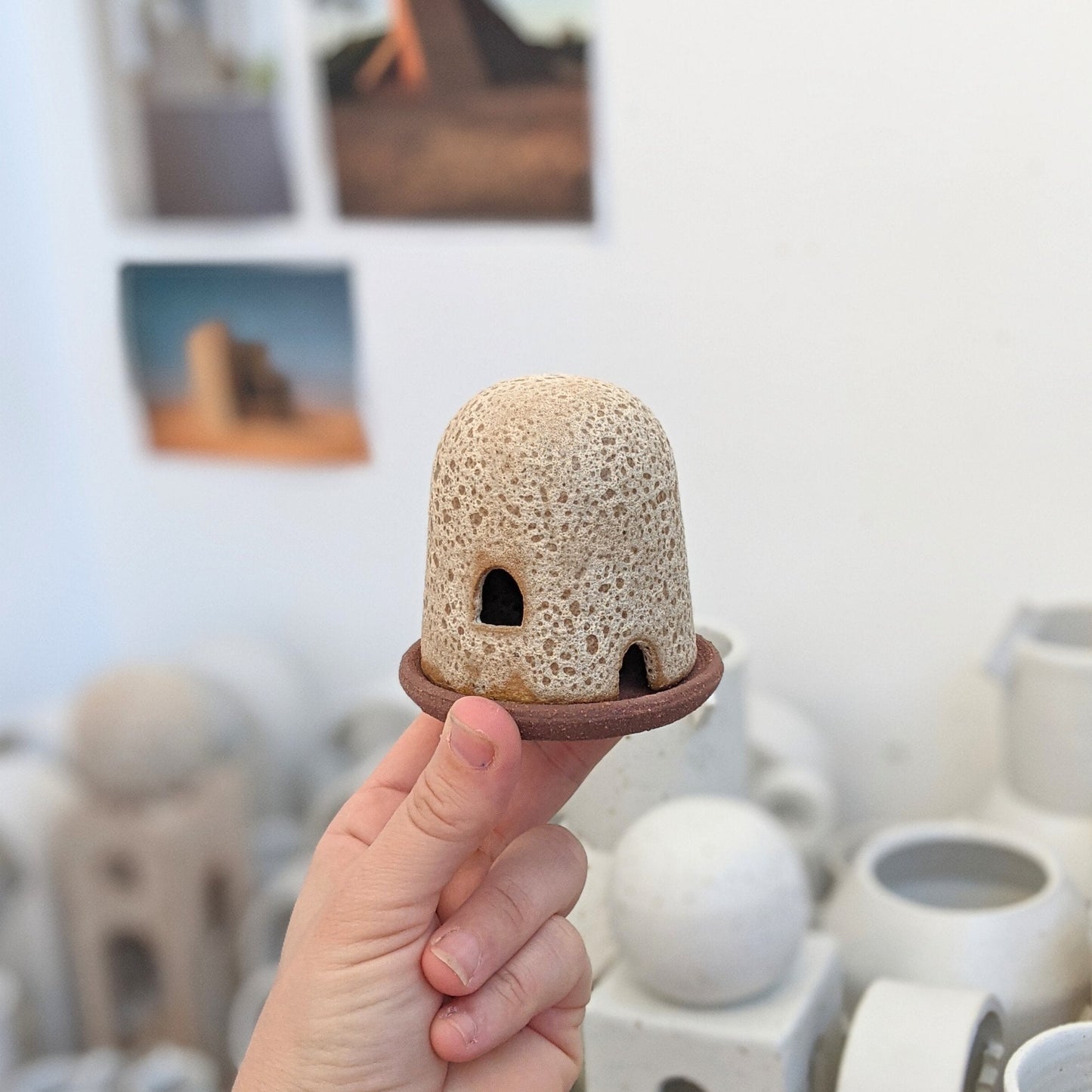 A hand holds a rust lava dome burner with ceramics in the background. The dome burner has a smooth dome top with hand carved windows and sits on a dish.