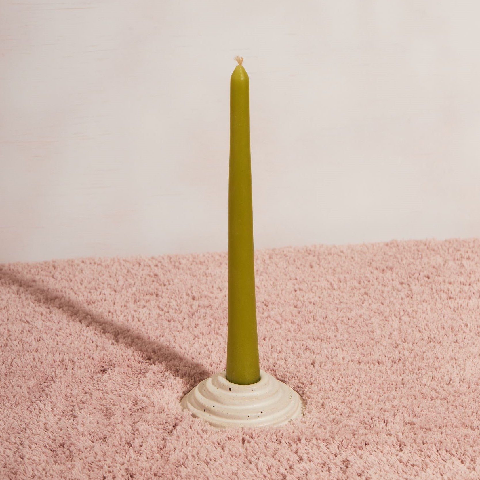 One circular candle holder in speckled white glaze sitting on pink fluffy fabric and a white background. The candle holder is holding one apple coloured beeswax candle