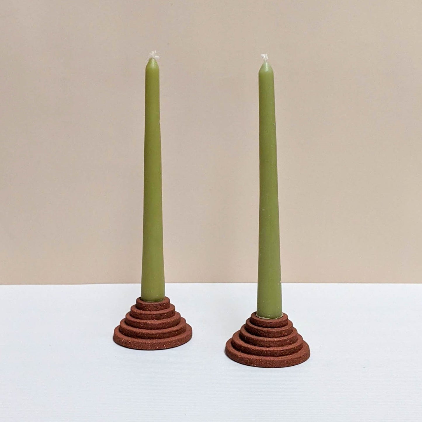 Two circular ceramic candle holders in terracotta. Each candle features steps leading up to the candle. Both of the candle holders have green unlit candles and are sitting on a white and cream backdrop.