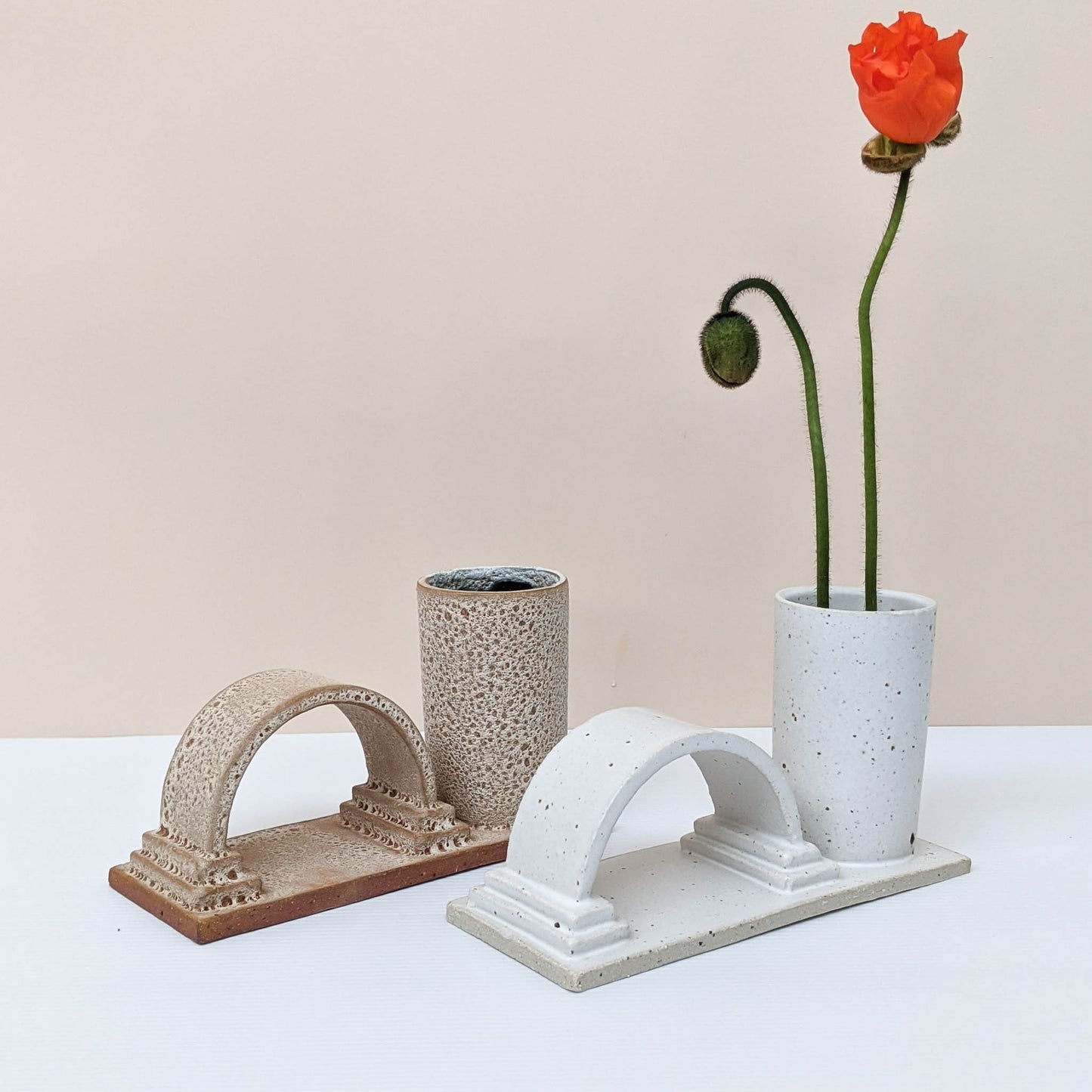 Two arch budvases handmade by Oh Hey Grace in Melbourne photographed on a white and cream background. The budvase on the left is finished in a textural lava glaze and the piece on the right is finished in a speckled cream glaze and has two poppies sitting in the vase.