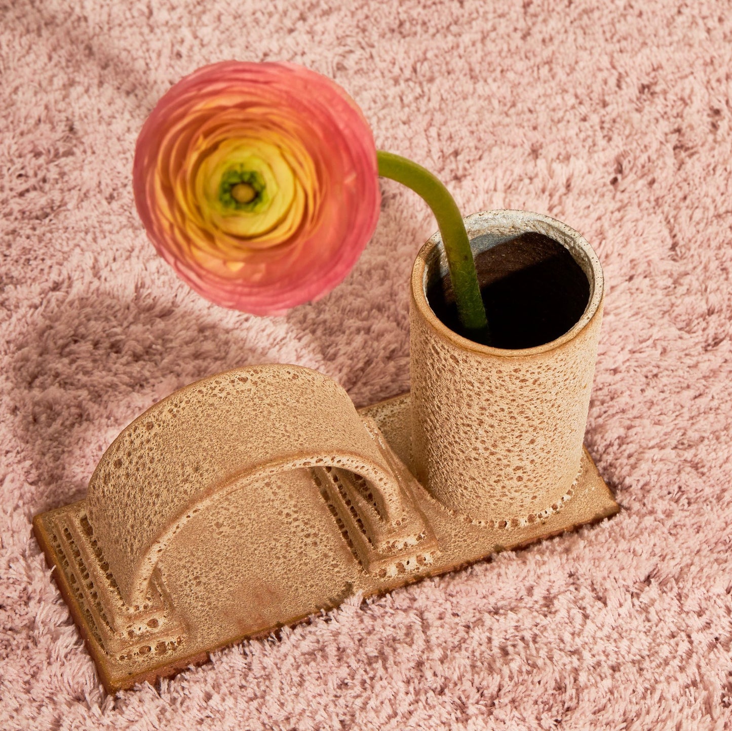 An arched budvase sits on a fluffy tablecloth with a white background. The budvase has a arch and a cylindrical vase on a rectangle base, finished in a rust lava glaze and has one flower sitting in the vase.