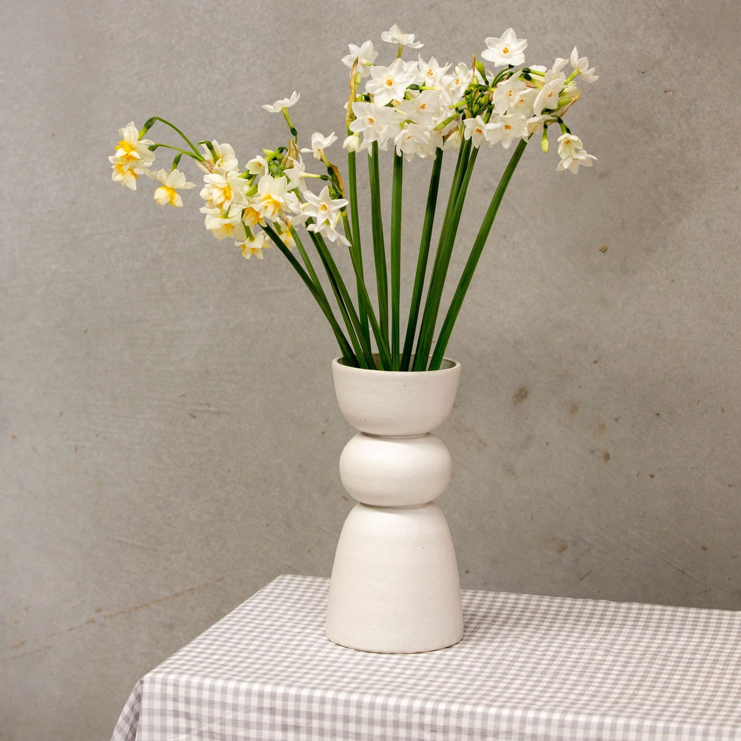 A handmade white vase with three distinct shapes stacked on each other holds white and yellow flowers and sits on a gingham tablecloth. 