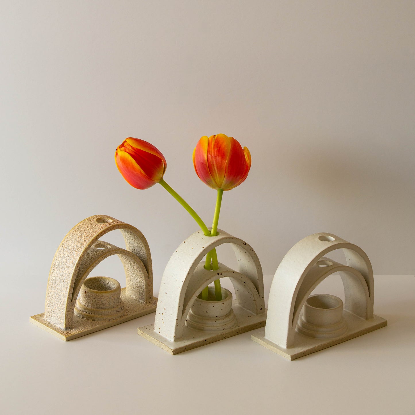Three handmade ceramic budvases on a white background. The budvases are in three different colourways. From left to right: Lunar, speckled cream, and white. The middle speckled cream budvase is holding two orange tulips.