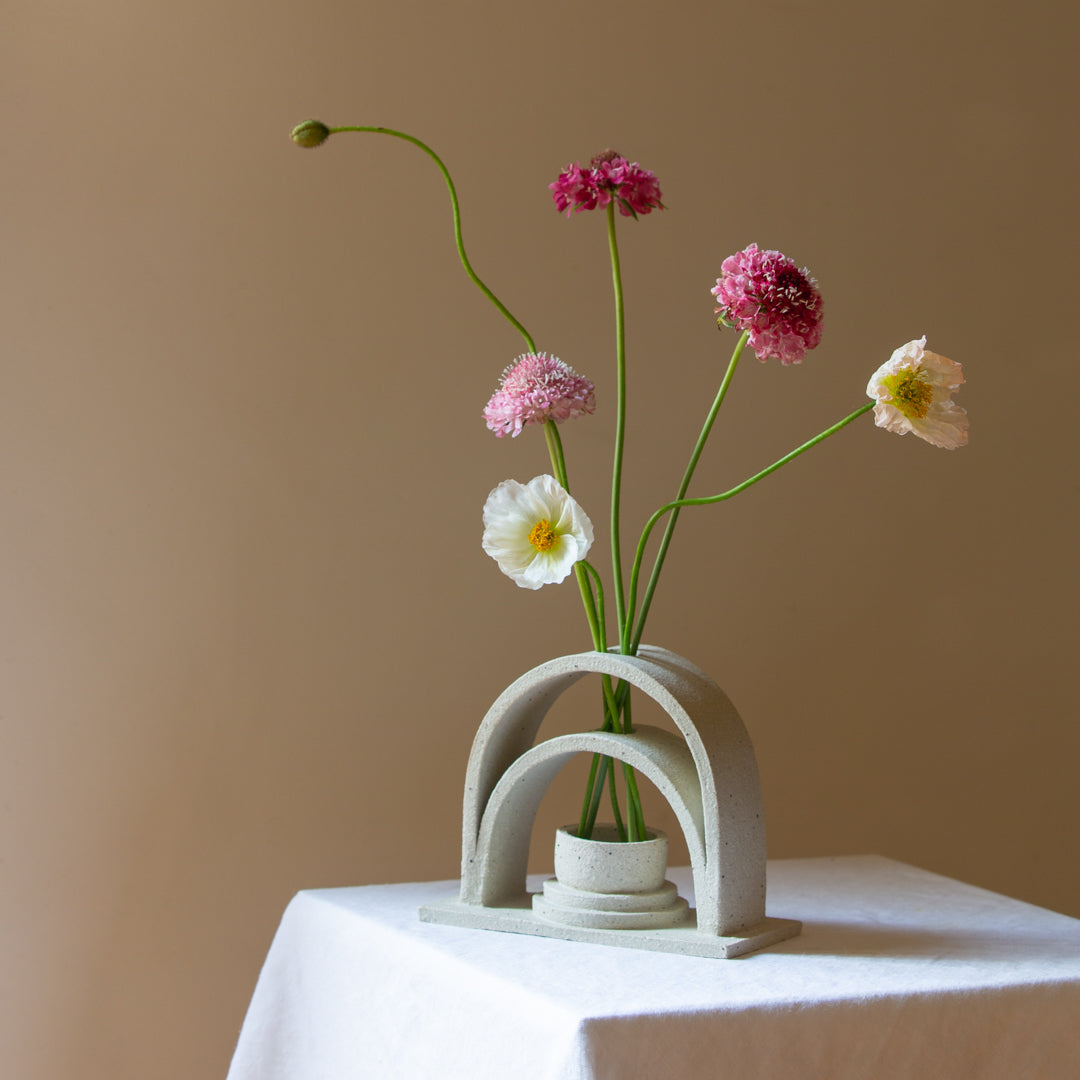 A handmade ceramic budvase sits on a white plinth. The budvase is in a sand colourway and has been left unglazed. The budvase holds a collection of pink, white and peach coloured flowers and unopened poppies.