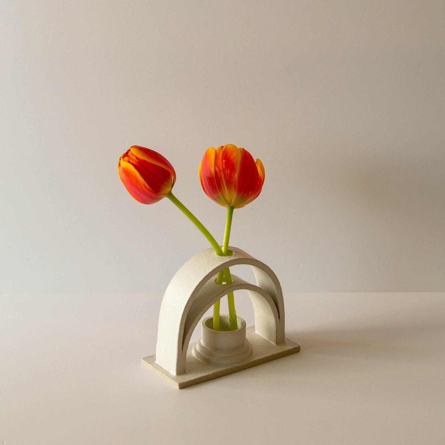 A handmade ceramic budvase on a white background. The budvase is finished in a white glaze and is is holding two orange tulips.