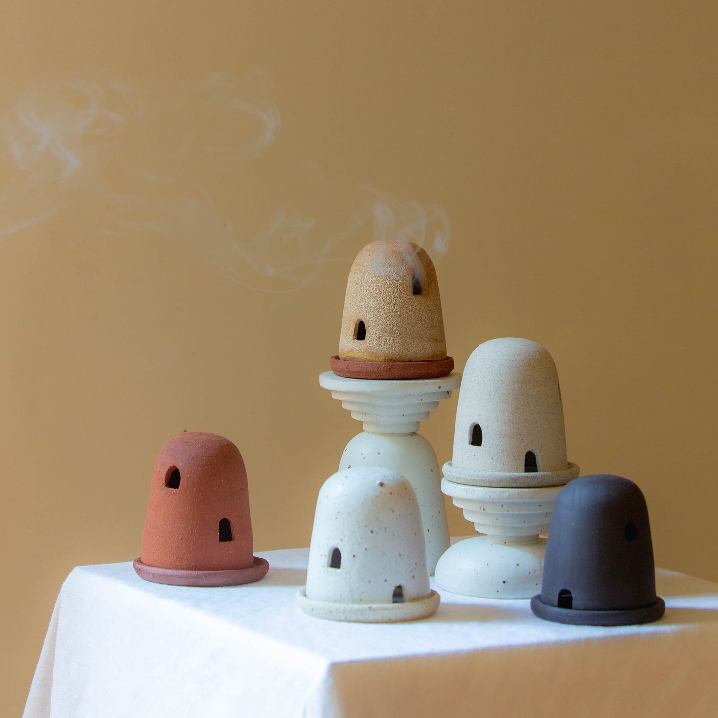 Five dome burners in terracotta, rust lava, speckled cream, grey, and black. Smoke is billowing out of the highest dome from a lit incense cone underneath. The dome burners have a smooth dome top with hand carved windows and sit on a dish.