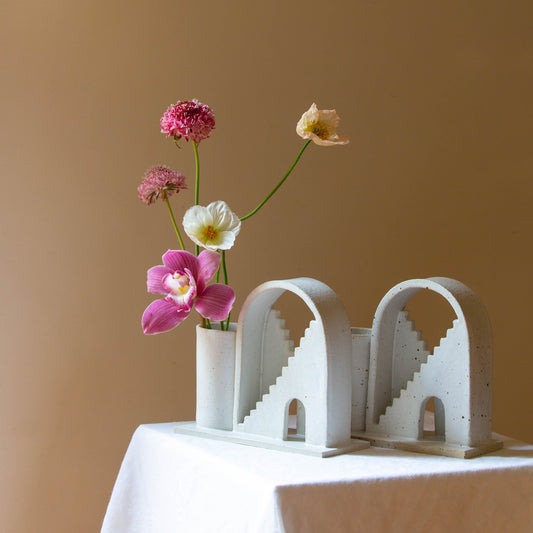 Two handmade ceramic vases with arched details and two mirrored staircases on each vase. The vase on the left is finished in a white glaze and has five flowers in the vase. The piece on the right has a speckled cream glaze. The budvases sits on a white plinth and is photographed on a cream background.