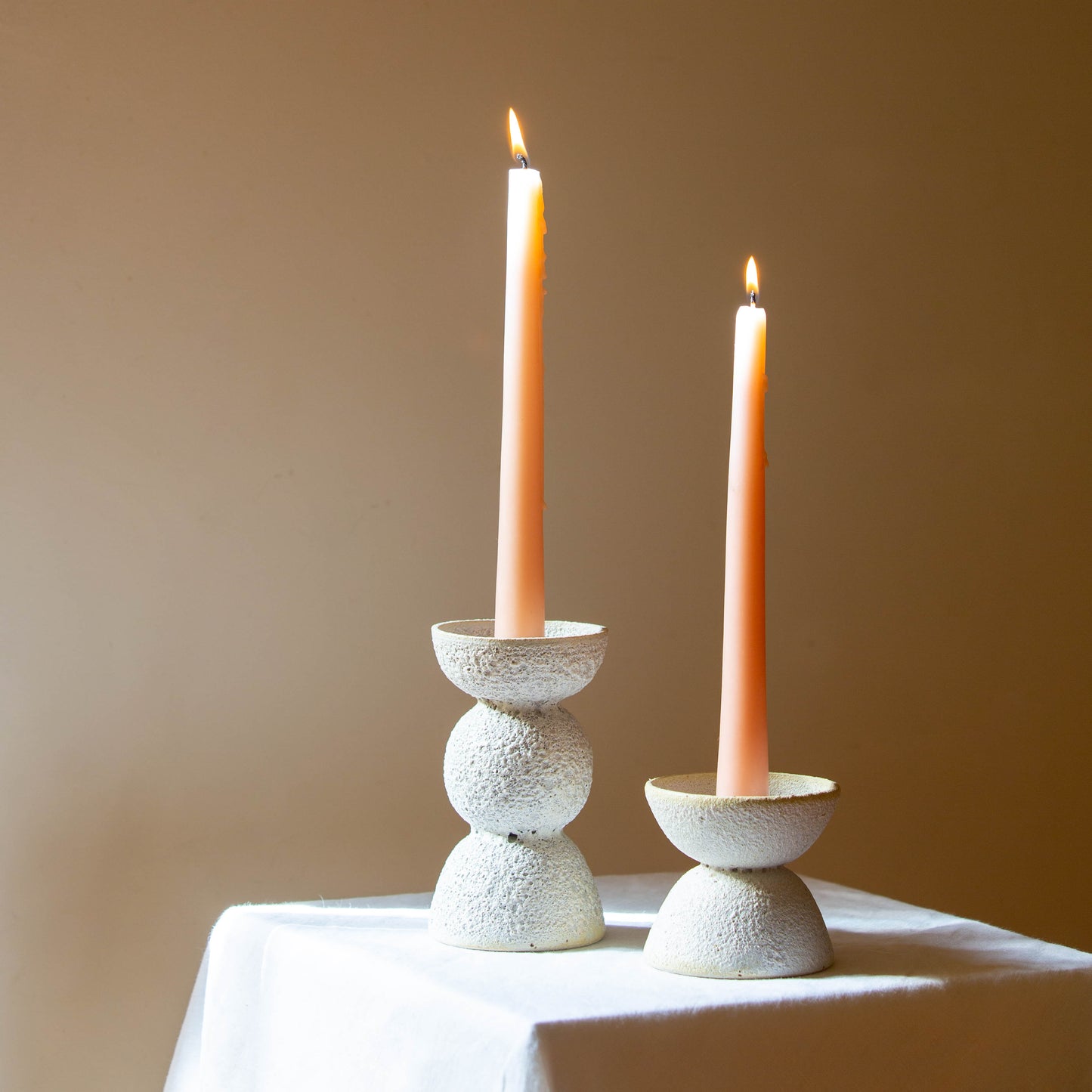 A tall and short candle holder hold lit candles. The candles are a peach colour by 'this is someday'