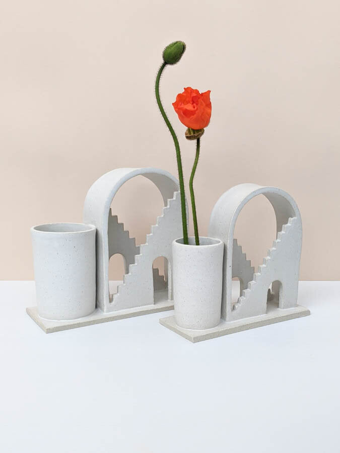 Two handmade ceramic vases with arched details and two mirrored staircases on each. The piece on the left is a large size, and the right is a regular size. The budvases are glazed in white. The budvase on the right hand side has two poppies inside the vase.  