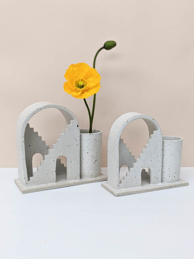 Two handmade ceramic vases with arched details and two mirrored staircases on each. The piece on the left is a large size, and the right is a regular size. The budvases are glazed in speckled white. The budvase on the left hand side has two poppies inside the vase.  
