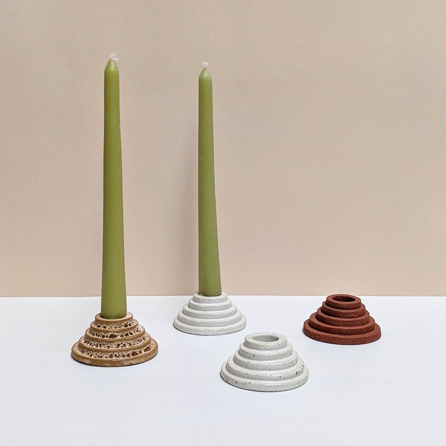 4  circular ceramic candle holders in rust lava, white, speckled cream, and terracotta colours.  Each candle features steps leading up to the candle. Two of the candle holders have green unlit candles and are sitting on a white and cream backdrop. 