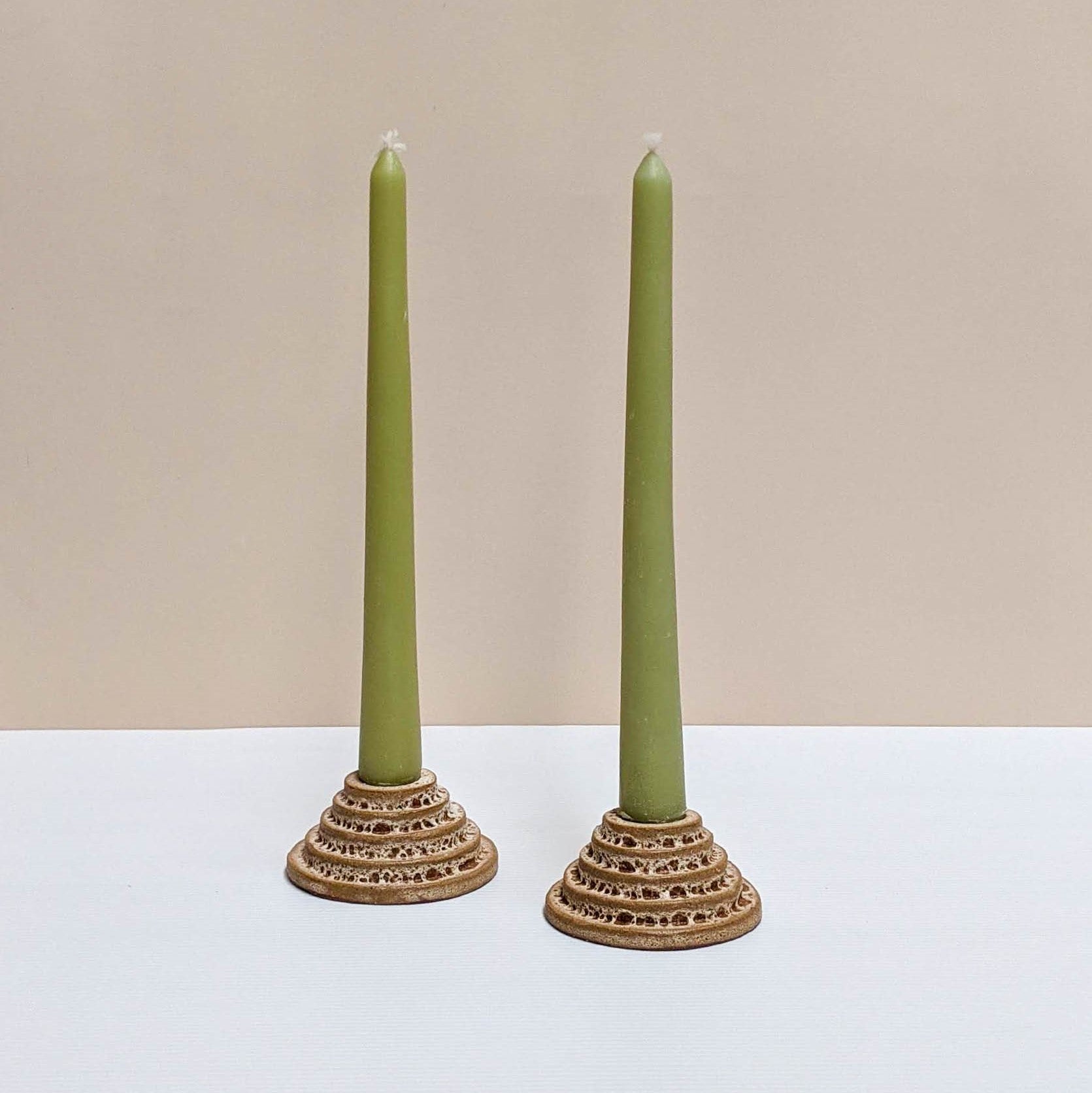 Two circular ceramic candle holders in rust lava glaze.  Each candle features steps leading up to the candle. Both of the candle holders have green unlit candles and are sitting on a white and cream backdrop. 