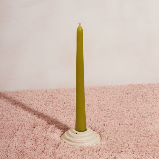 A circular ceramic candle holder featuring steps leading up to the candle. This candle holder has a green unlit candle and is sitting on a pink fluffy fabric with a white backdrop.