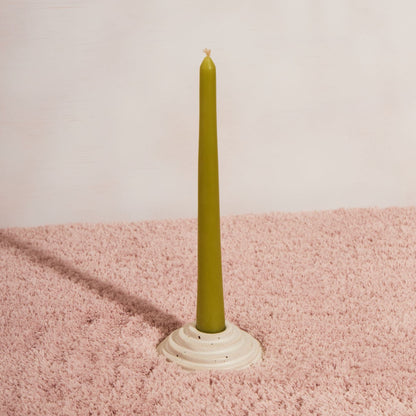 One circular candle holder in speckled white glaze sitting on pink fluffy fabric and a white background. The candle holder is holding one apple coloured beeswax candle 