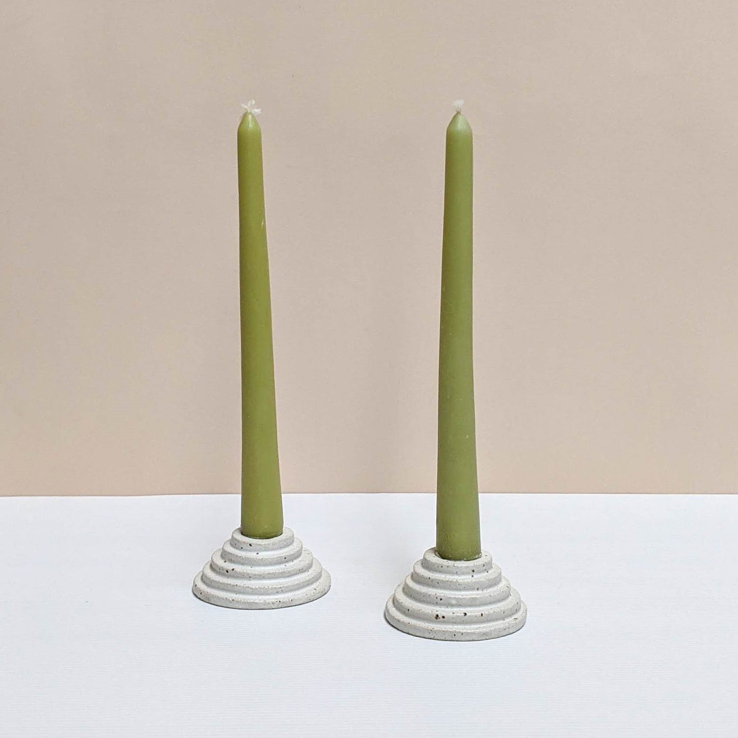 Two circular ceramic candle holders in speckled cream glaze.  Each candle features steps leading up to the candle. Both of the candle holders have green unlit candles and are sitting on a white and cream backdrop. 