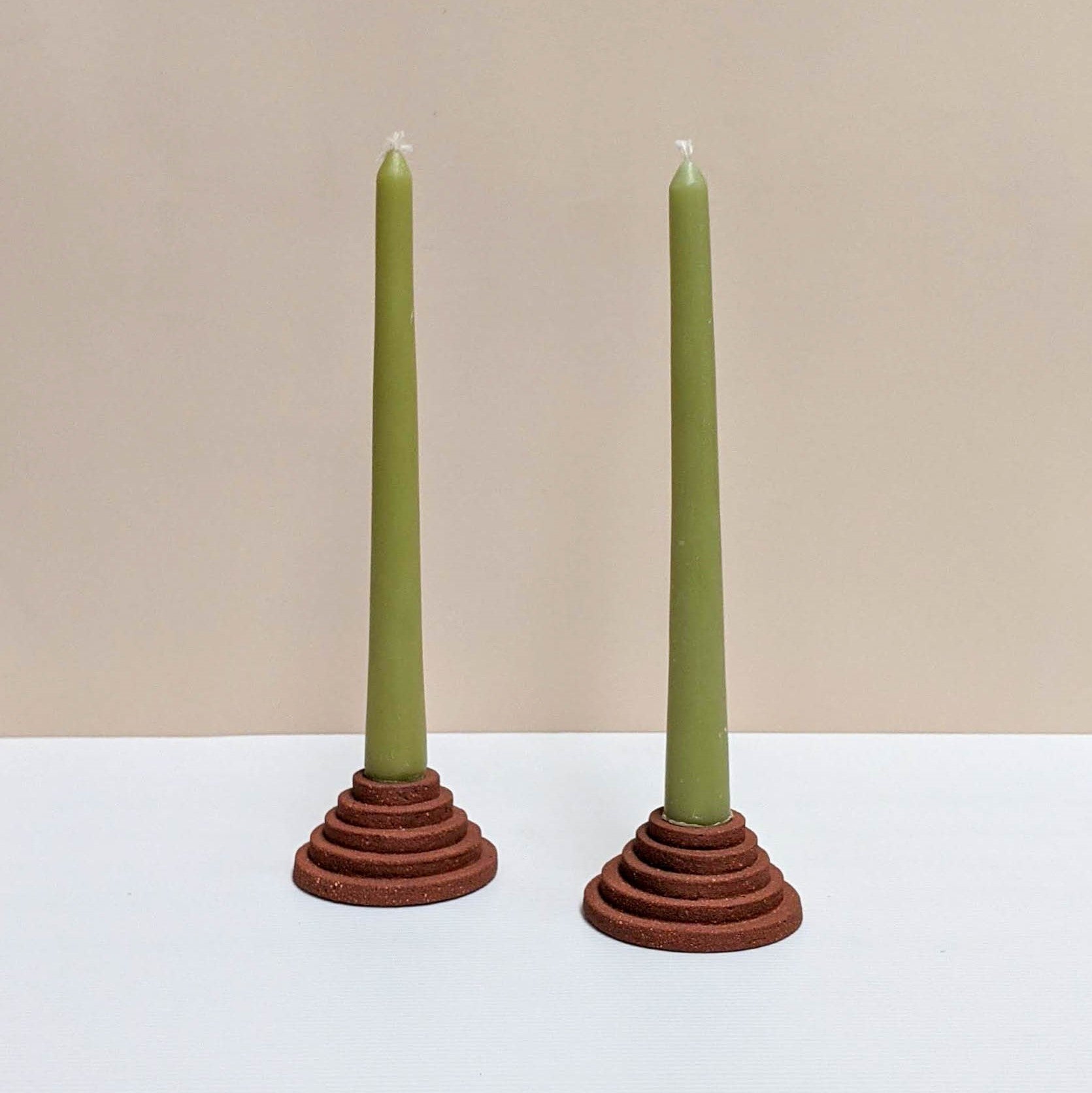 Two circular ceramic candle holders in terracotta.  Each candle features steps leading up to the candle. Both of the candle holders have green unlit candles and are sitting on a white and cream backdrop. 
