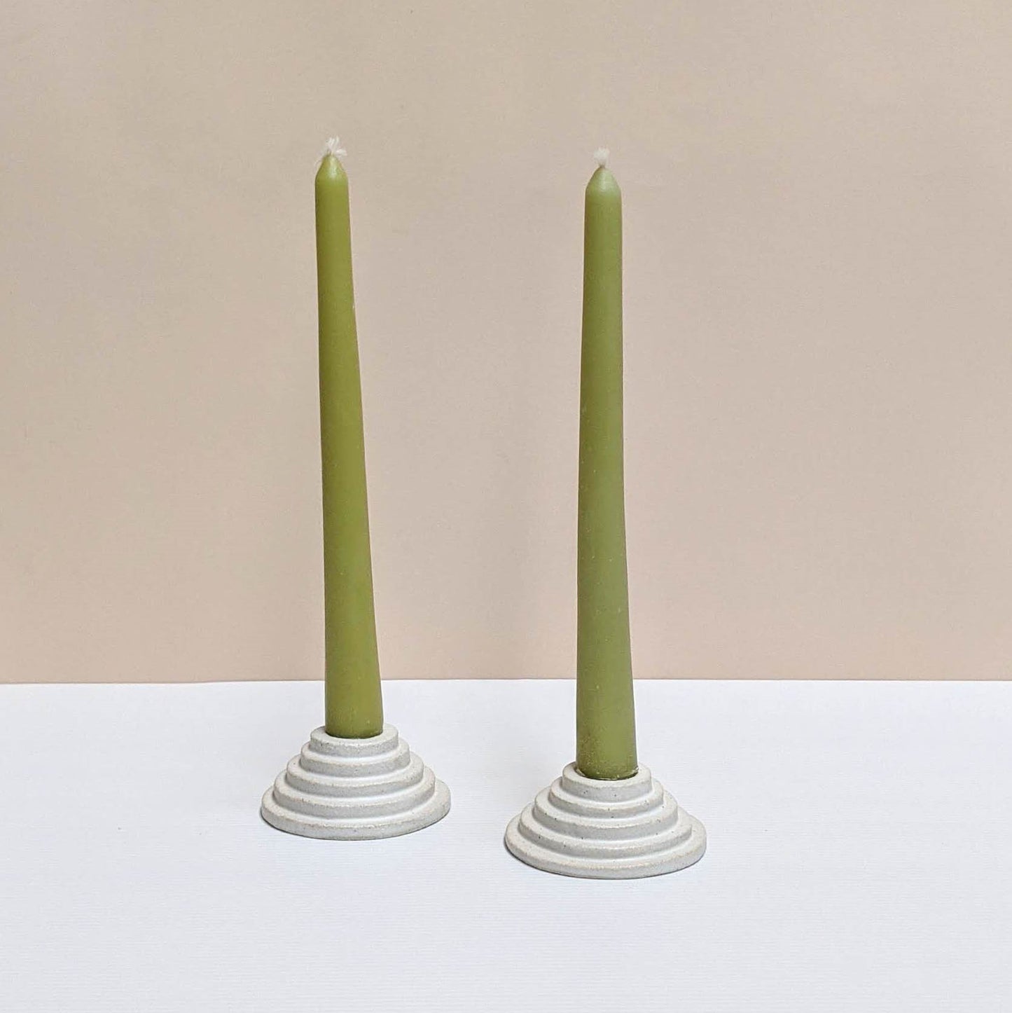 Two circular ceramic candle holders in white glaze.  Each candle features steps leading up to the candle. Both of the candle holders have green unlit candles and are sitting on a white and cream backdrop. 
