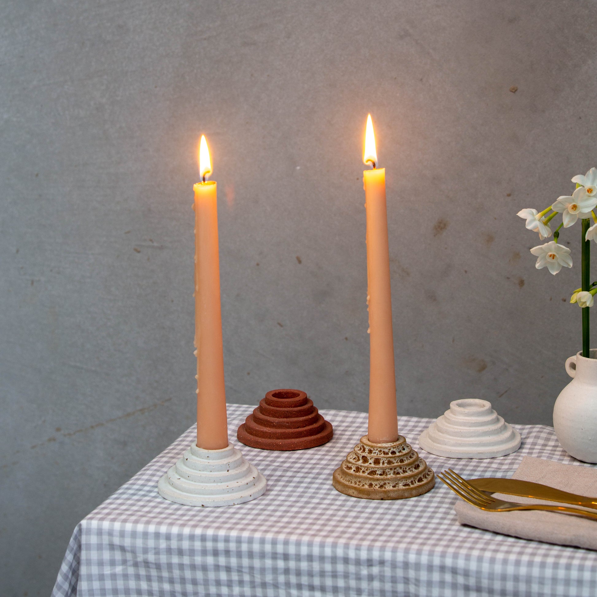 A circular ceramic candle holder featuring steps leading up to the candle. These candle holders have peach candles burning and are sitting on a gingham tablecloth with a vase and flowers, and gold cutlery on a linen napkin.