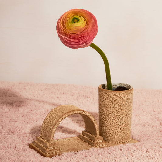 An arched budvase sits on a fluffy tablecloth with a white background. The budvase has a arch and a cylindrical vase on a rectangle base, finished in a rust lava glaze and has one flower sitting in the vase.
