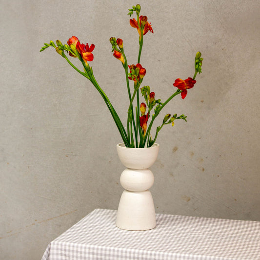 A white vase with three distinct shapes stacked on each other holds red flowers and sits on a gingham tablecloth. 