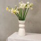 A handmade white vase with three distinct shapes stacked on each other holds white and yellow flowers and sits on a gingham tablecloth. 