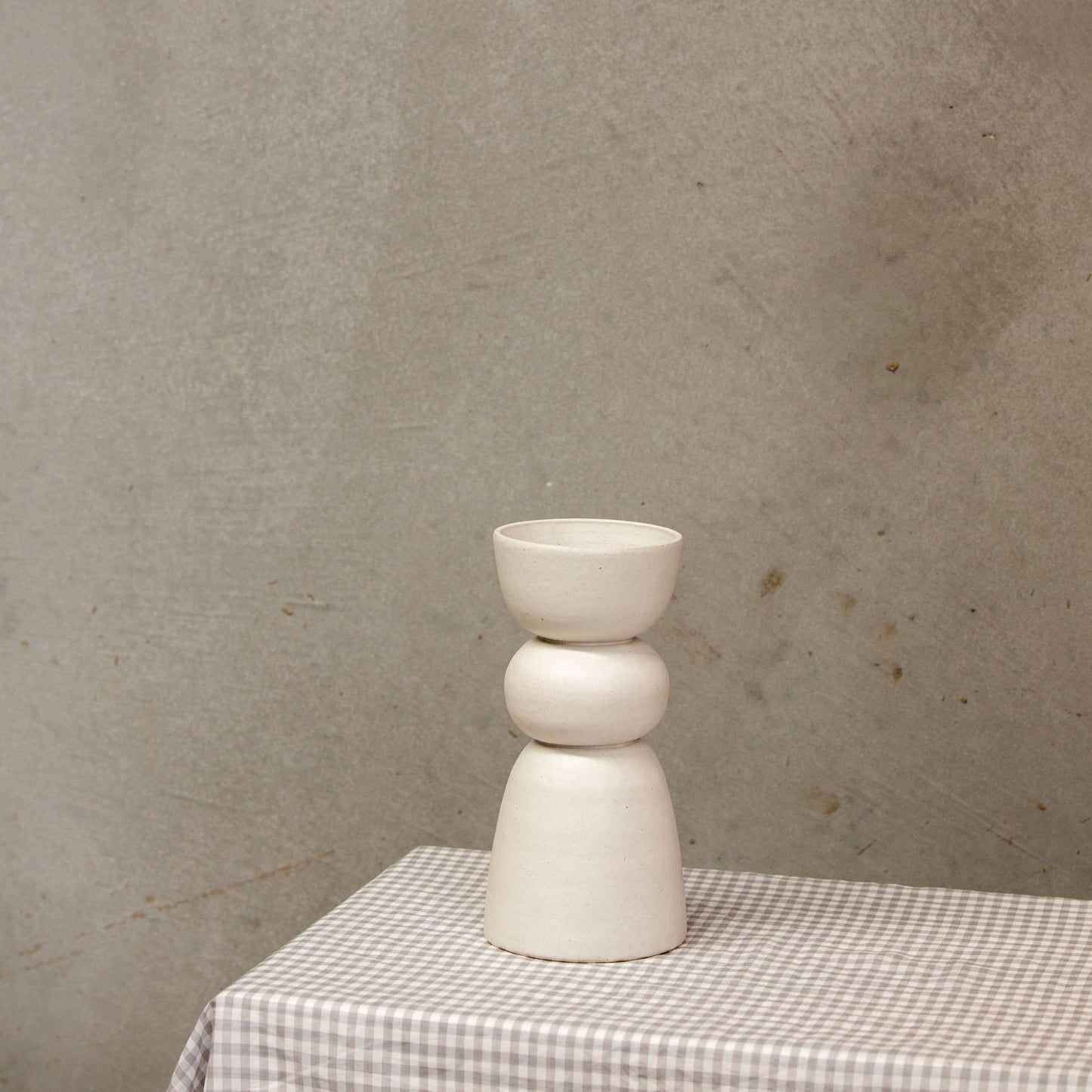 A white vase with three distinct shapes stacked on each other and sits on a gingham tablecloth. 
