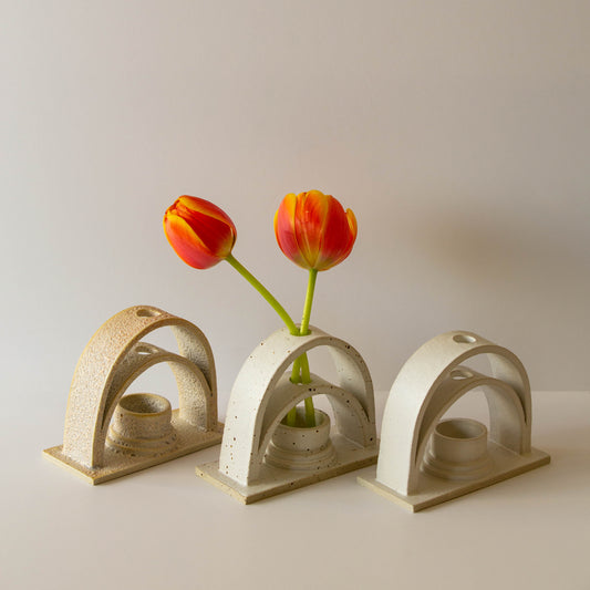 Three handmade ceramic budvases on a white background. The budvases are in three different colourways. From left to right: Lunar, speckled cream, and white. The middle speckled cream budvase is holding two orange tulips. 