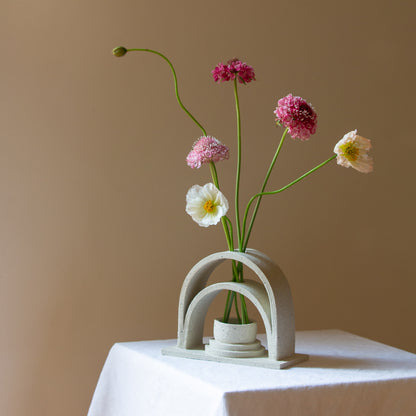 A handmade ceramic budvase sits on a white plinth. The budvase is in a sand colourway and has been left unglazed. The budvase holds a collection of pink, white and peach coloured flowers and unopened poppies.