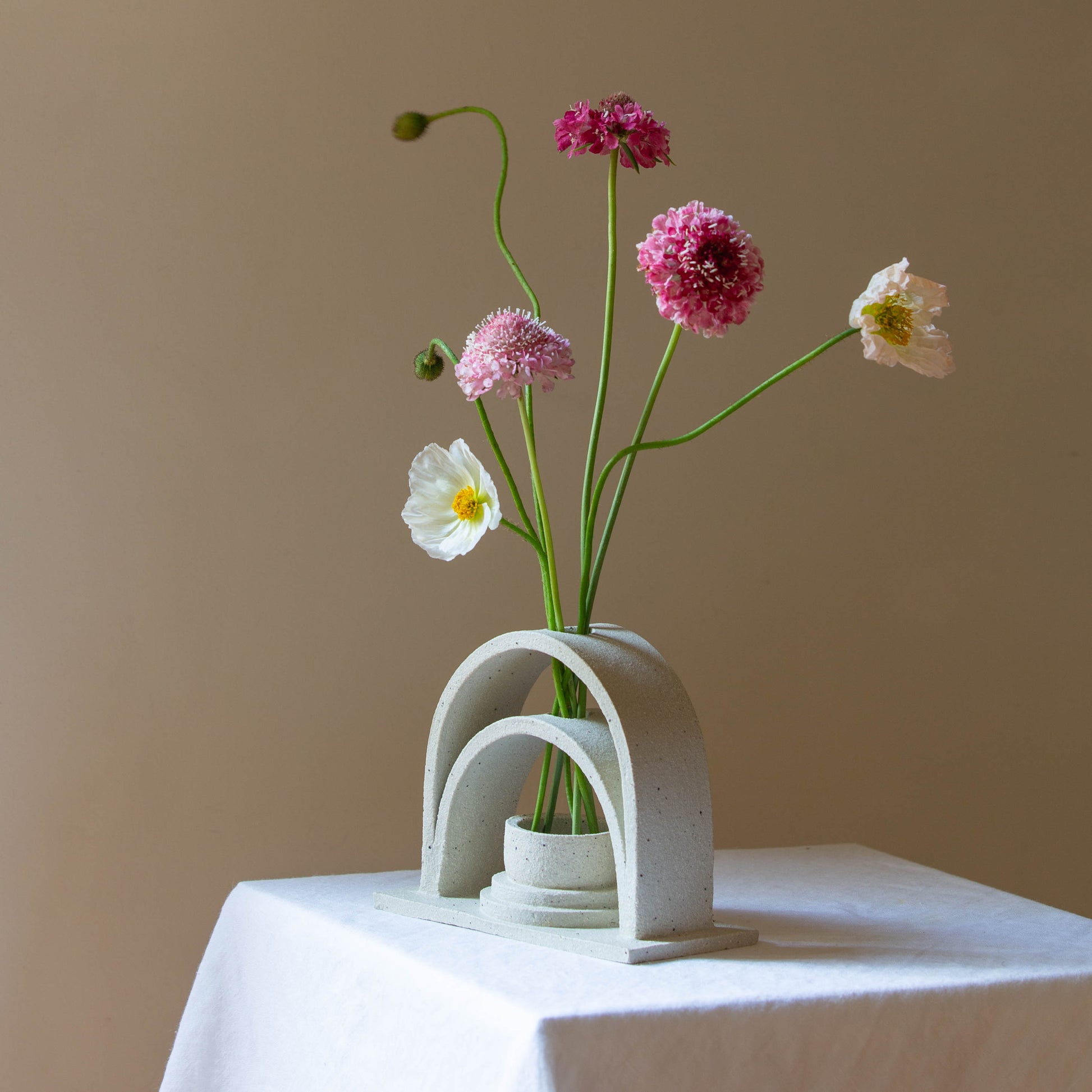 A handmade ceramic budvase sits on a white plinth. The budvase is in a sand colourway and has been left unglazed. The budvase holds a collection of pink, white and peach coloured flowers and unopened poppies.