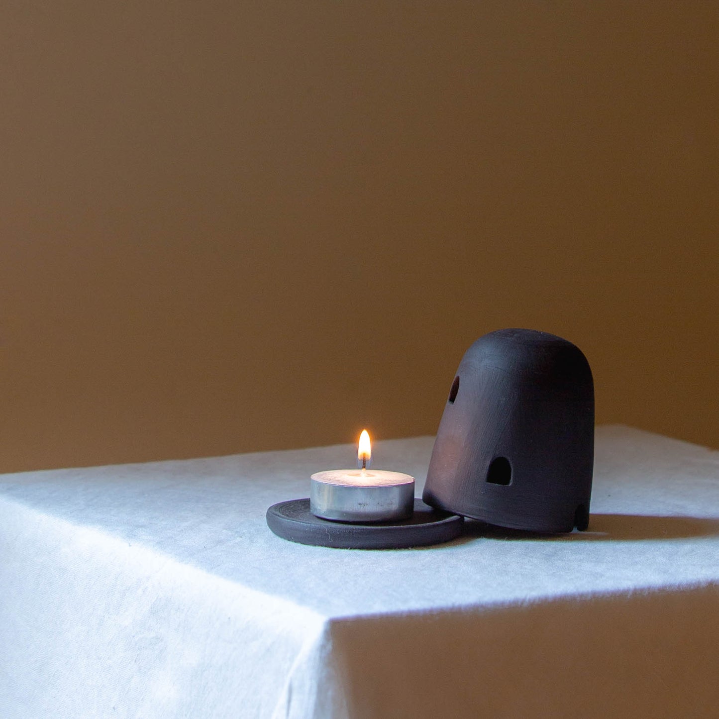 Black Dome Burner sitting on a white plinth with a lit candle. The dome burner has a smooth dome top with hand carved windows and sits on a dish.