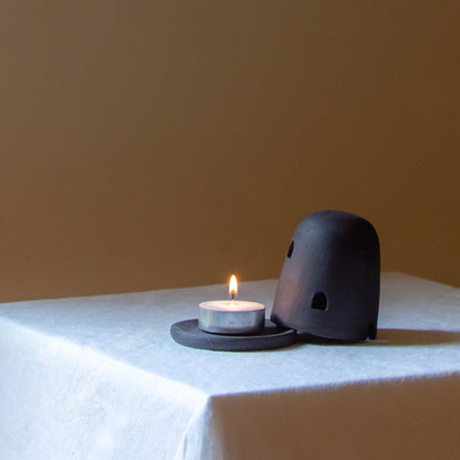 Black Dome Burner sitting on a white plinth with a lit candle. The dome burner has a smooth dome top with hand carved windows and sits on a dish.