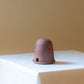 A Dome Burner in terracotta sitting on a white plinth . The dome burner has a smooth dome top with hand carved windows and sits on a dish. A lit candle sits inside making the windows glow.