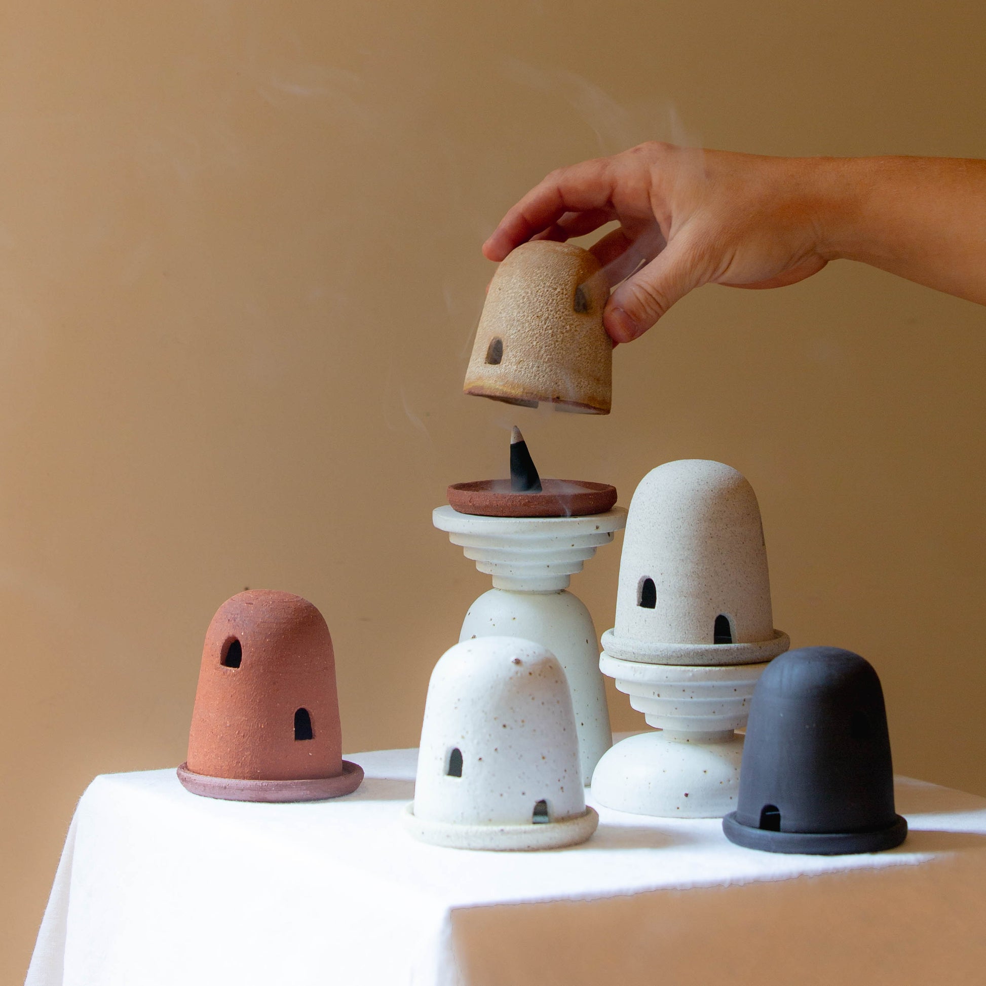 Five dome burners in terracotta, rust lava, speckled cream, grey, and black. A hand carefully holds one dome and smoke is billowing out while a incense cone burns on the dish. The dome burners have a smooth dome top with hand carved windows and sit on a dish.
