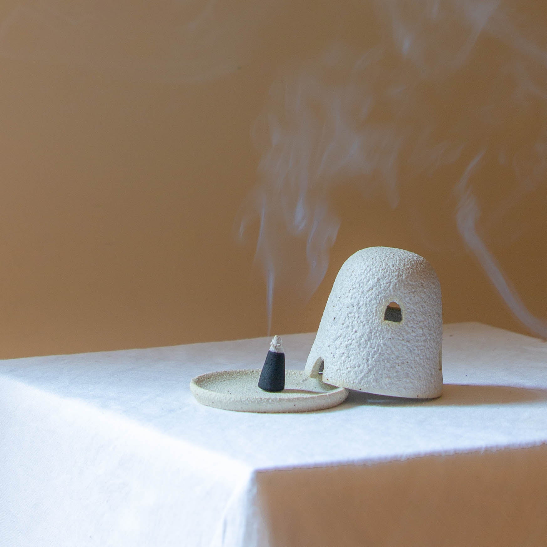 A Dome Burner in lunar glaze sitting on a white plinth with a lit incense cone which is billowing smoke. The dome burner has a smooth dome top with hand carved windows and sits on a dish.