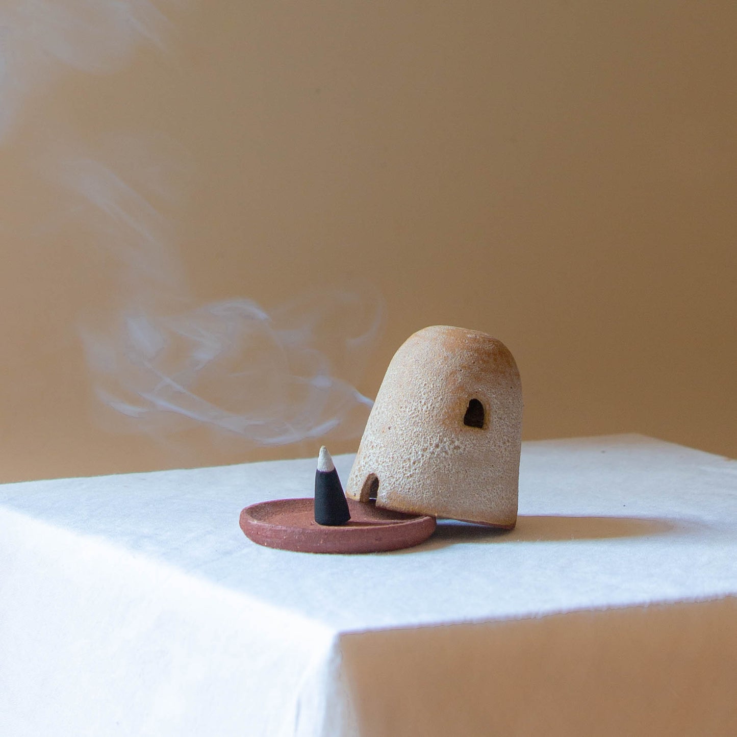 A Dome Burner in rust lava glaze sitting on a white plinth with a lit incense cone which is billowing smoke. The dome burner has a smooth dome top with hand carved windows and sits on a dish.