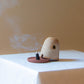 A Dome Burner in rust lava glaze sitting on a white plinth with a lit incense cone which is billowing smoke. The dome burner has a smooth dome top with hand carved windows and sits on a dish.