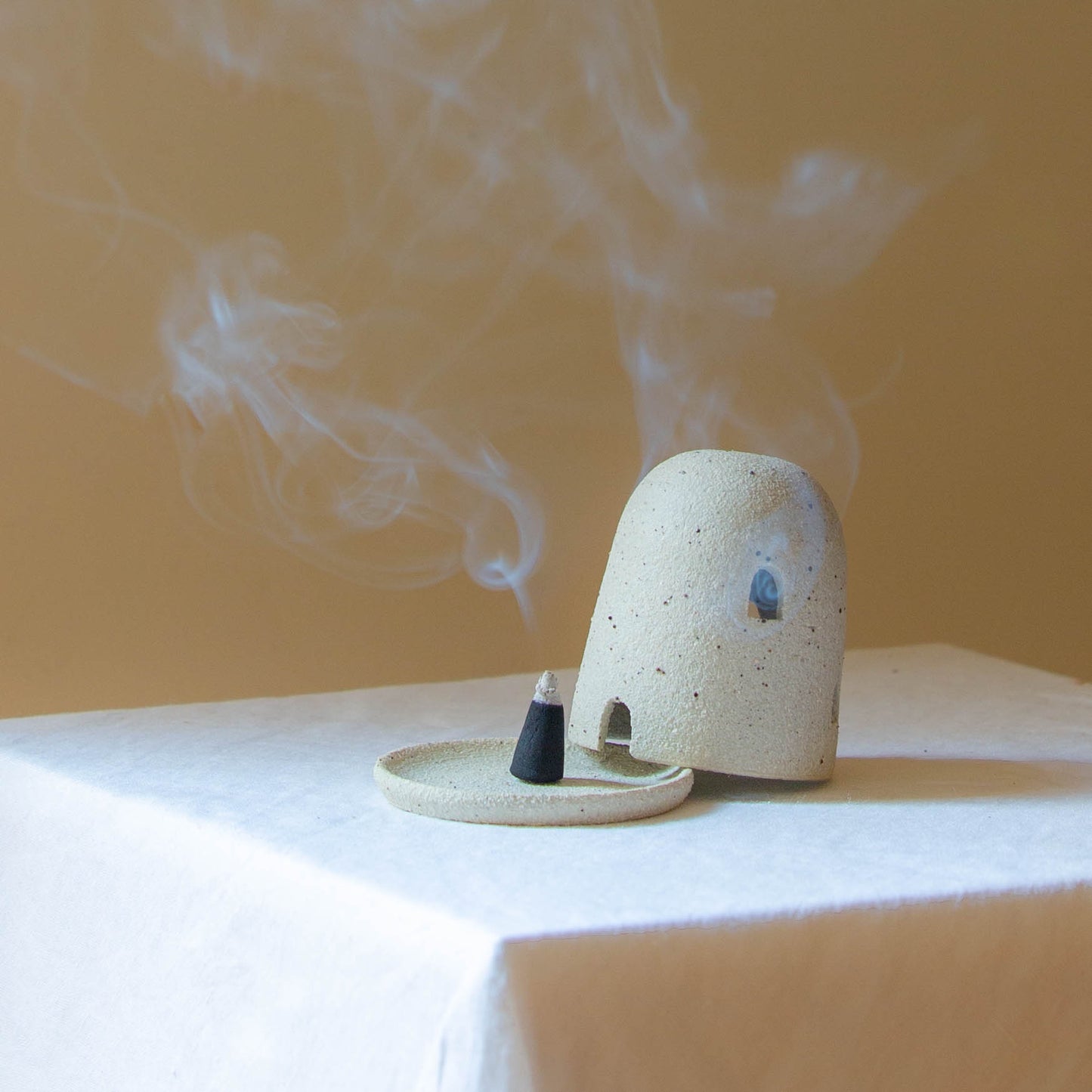 A Dome Burner in raw sand colour sitting on a white plinth with a lit incense cone which is billowing smoke. The dome burner has a smooth dome top with hand carved windows and sits on a dish.