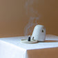 A Dome Burner in speckled cream glaze sitting on a white plinth with a lit incense cone which is billowing smoke. The dome burner has a smooth dome top with hand carved windows and sits on a dish.