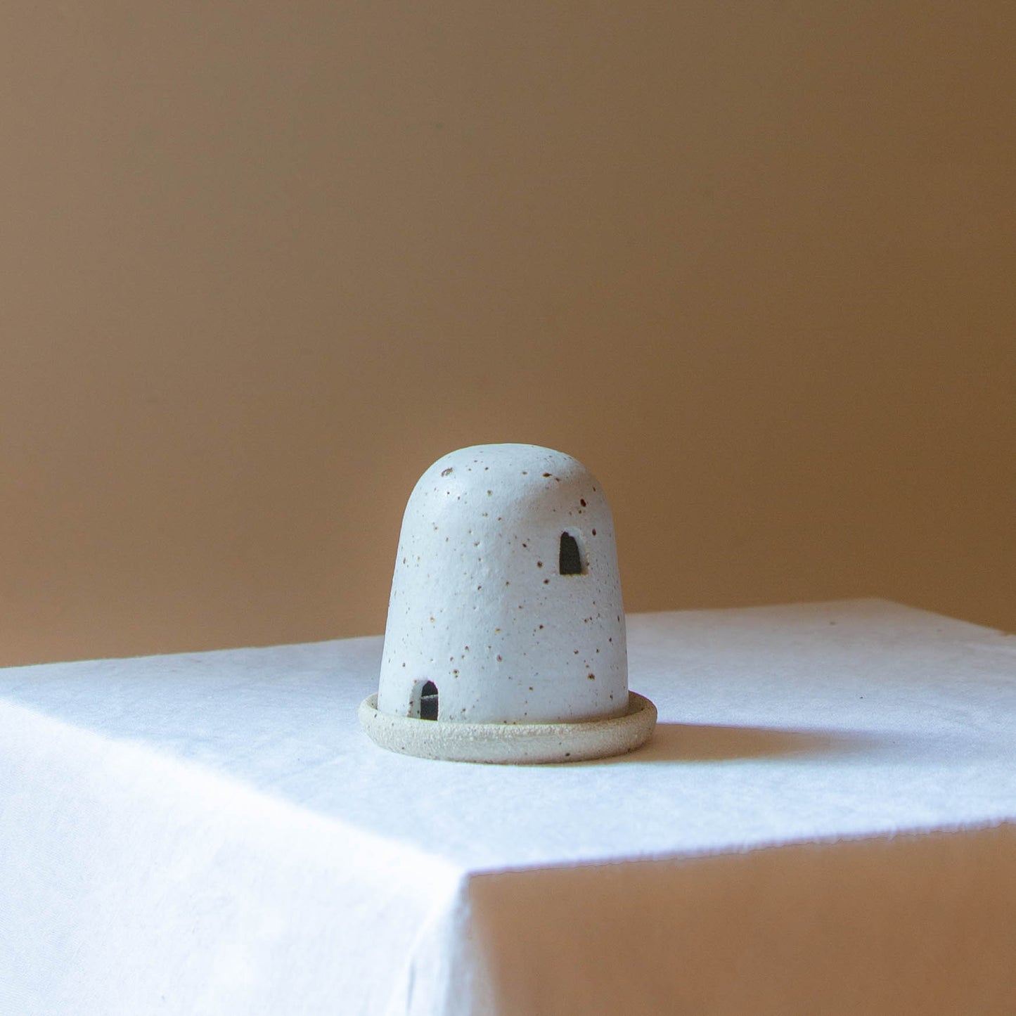 A Dome Burner in speckled cream glaze sitting on a white plinth. The dome burner has a smooth dome top with hand carved windows and sits on a dish.