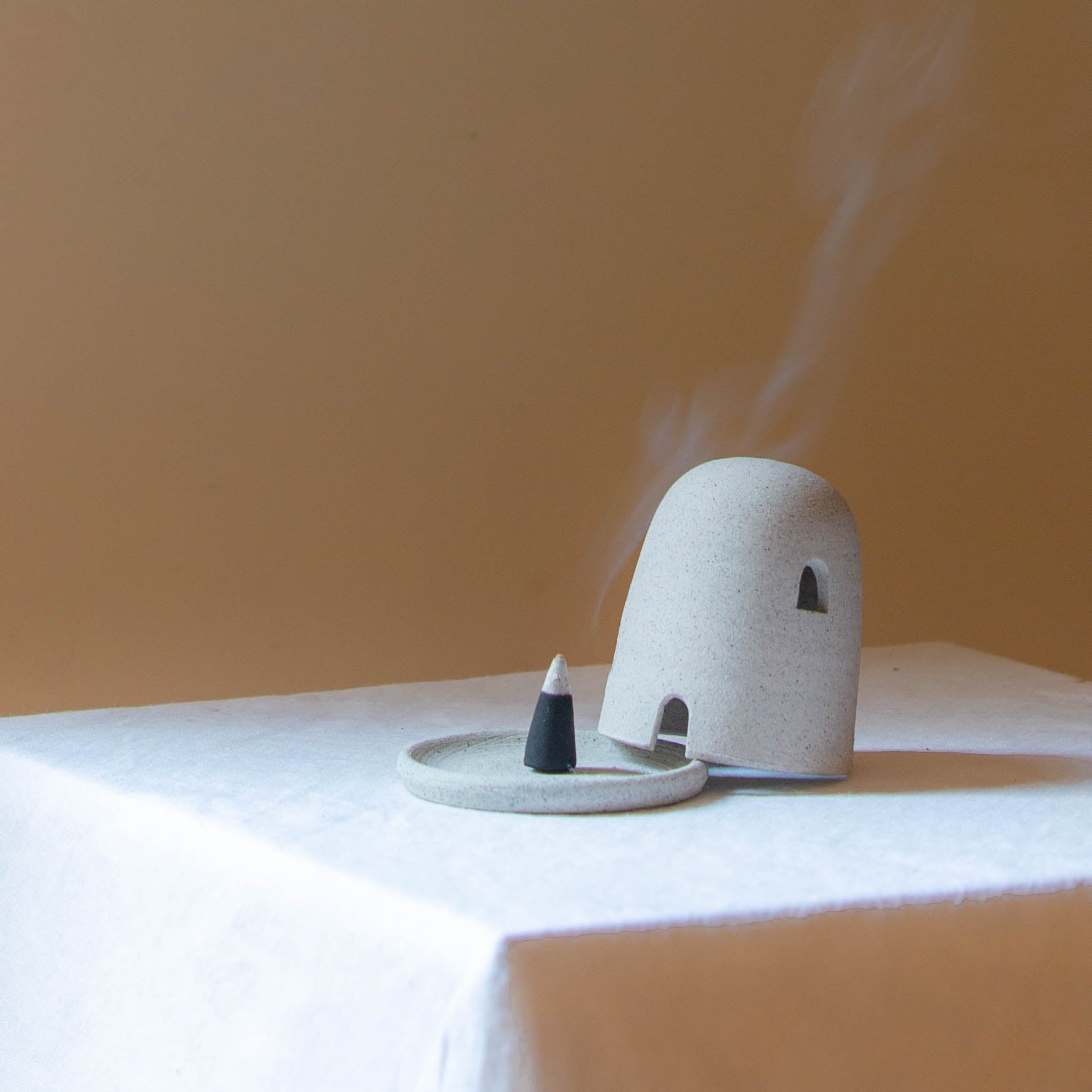 A Dome Burner in speckled grey colour sitting on a white plinth with a lit incense cone which is billowing smoke. The dome burner has a smooth dome top with hand carved windows and sits on a dish.