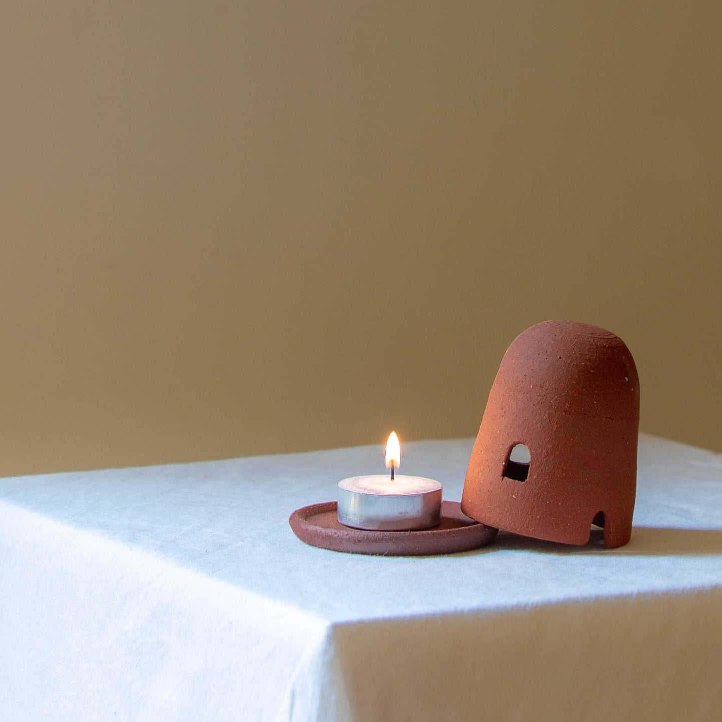 A Dome Burner in terracotta sitting on a white plinth with a lit incense cone which is billowing smoke. The dome burner has a smooth dome top with hand carved windows and sits on a dish.