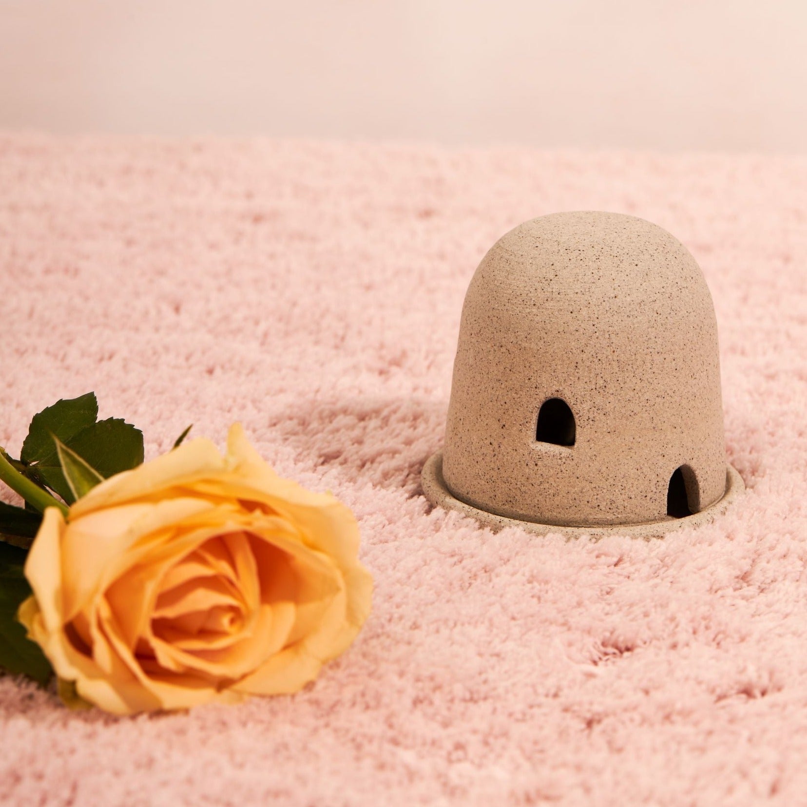 A grey dome burner sits on fluffy pink fabric with a yellow rose on the left. The dome burner has a smooth dome top with hand carved windows and sits on a dish.