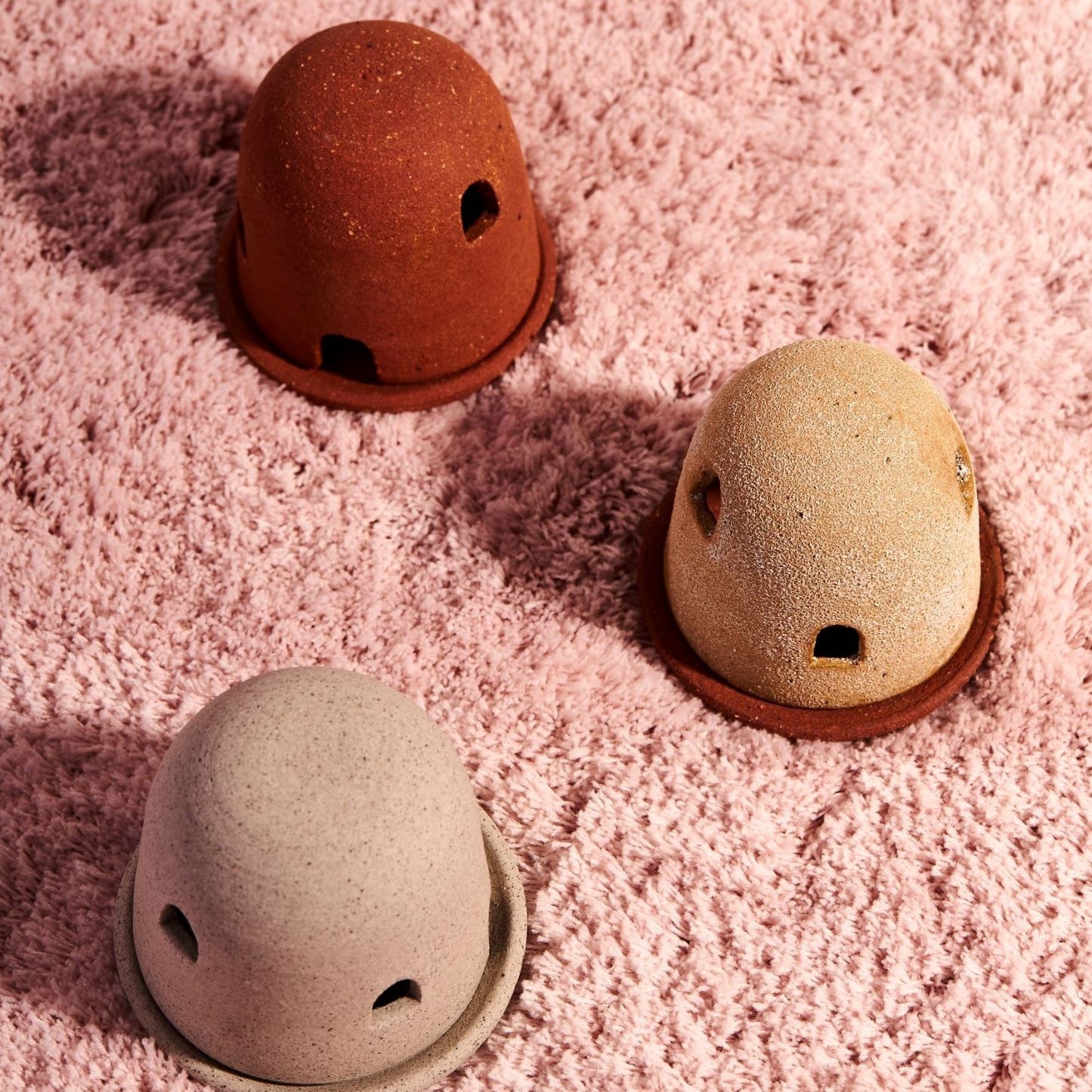 Three dome burners in grey, terracotta, and rust lava, sit on fluffy pink fabric. The dome burners have a smooth dome top with hand carved windows and sit on a dish.