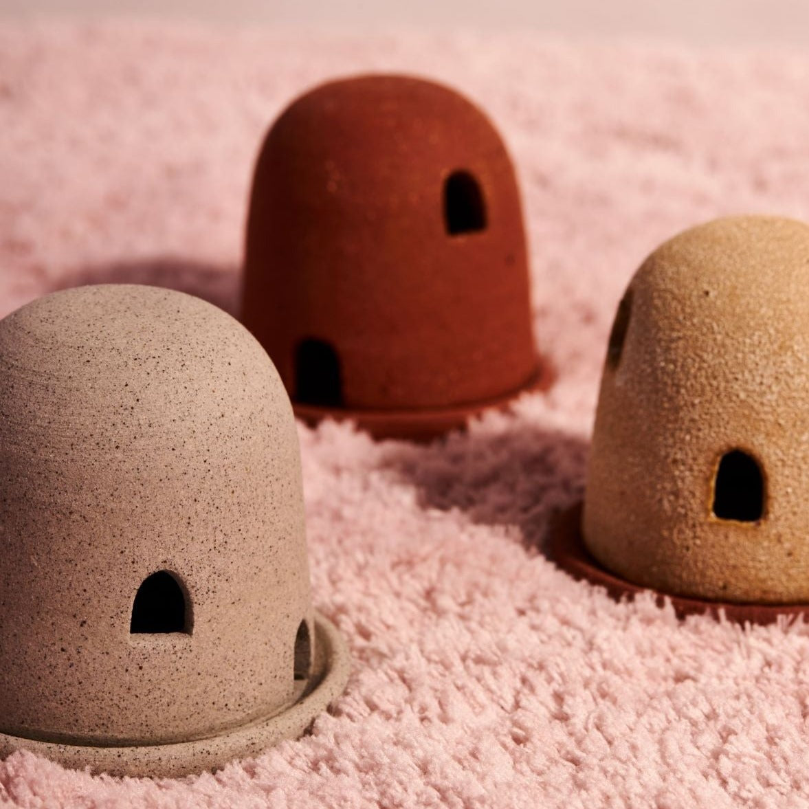 Three dome burners in grey, terracotta, and rust lava, sit on fluffy pink fabric. The dome burners have a smooth dome top with hand carved windows and sit on a dish.