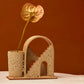 Handmade ceramic vase with arched detail and two mirrored staircases. The vase is finished in a rust lava glaze and has a single flower inside the vase. The budvase sits on a white plinth and is photographed on a terracotta background. 
