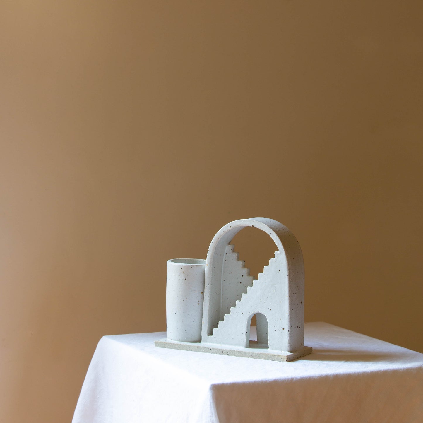 Handmade ceramic vase with arched detail and two mirrored staircases. The vase is finished in a speckled cream glaze. The budvase sits on a white plinth and is photographed on a cream background. 