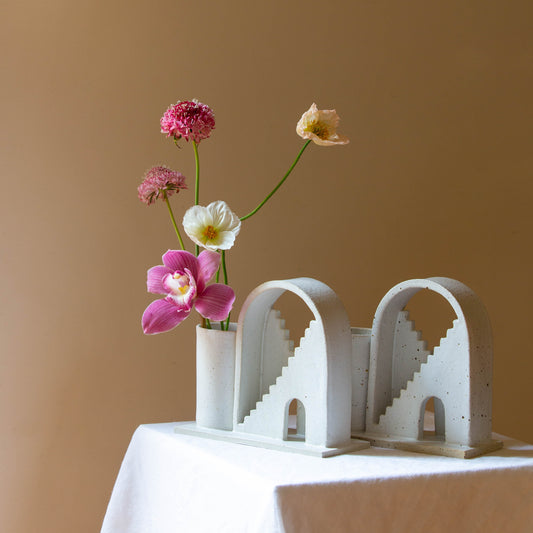 Two handmade ceramic vases with arched details and two mirrored staircases on each vase. The vase on the left is finished in a white glaze and has five flowers in the vase. The piece on the right has a speckled cream glaze. The budvases sits on a white plinth and is photographed on a cream background. 