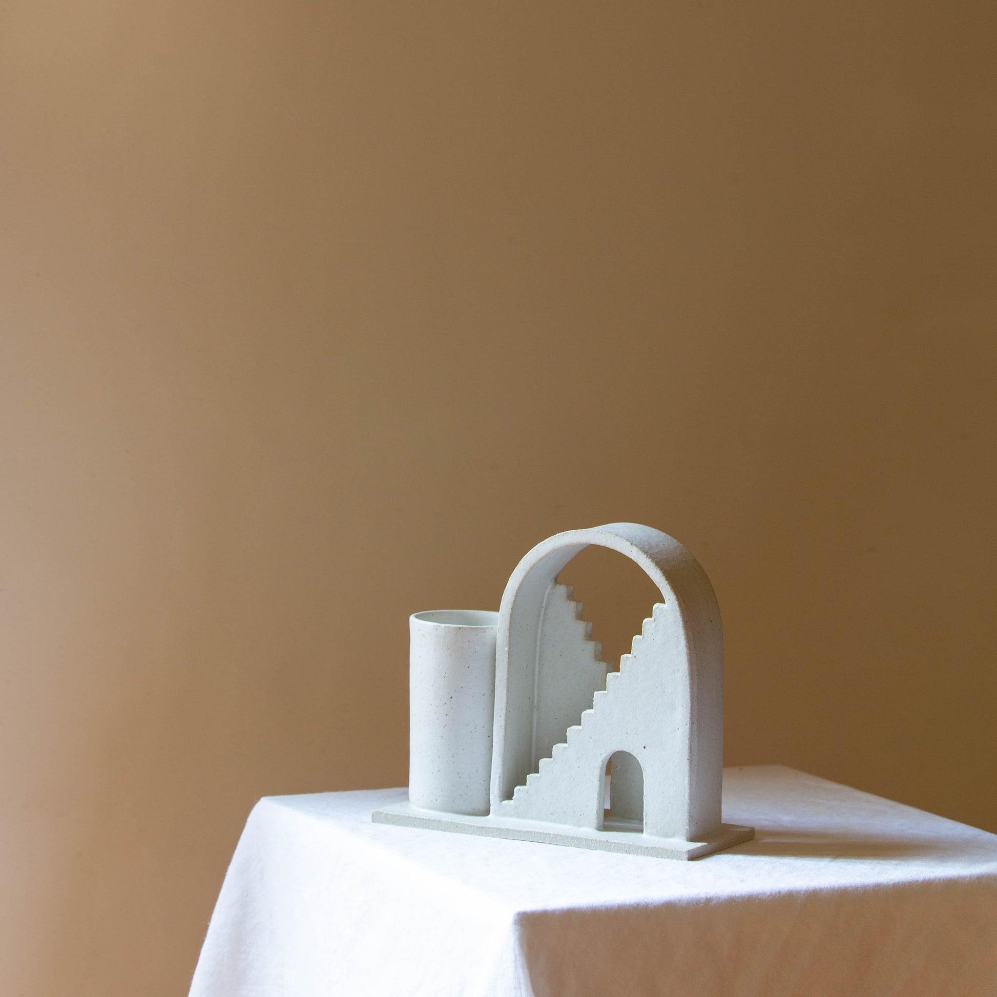 Handmade ceramic vase with arched detail and two mirrored staircases. The vase is finished in a white glaze. The budvase sits on a white plinth and is photographed on a cream background. 