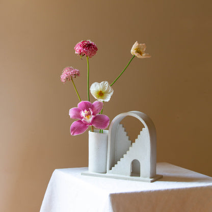 Handmade ceramic vase with arched detail and two mirrored staircases. The vase is finished in a white glaze and has five flowers in the vase. The budvase sits on a white plinth and is photographed on a cream background. 