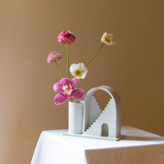 Handmade ceramic vase with arched detail and two mirrored staircases. The vase is finished in a white glaze and has five flowers in the vase. The budvase sits on a white plinth and is photographed on a cream background.